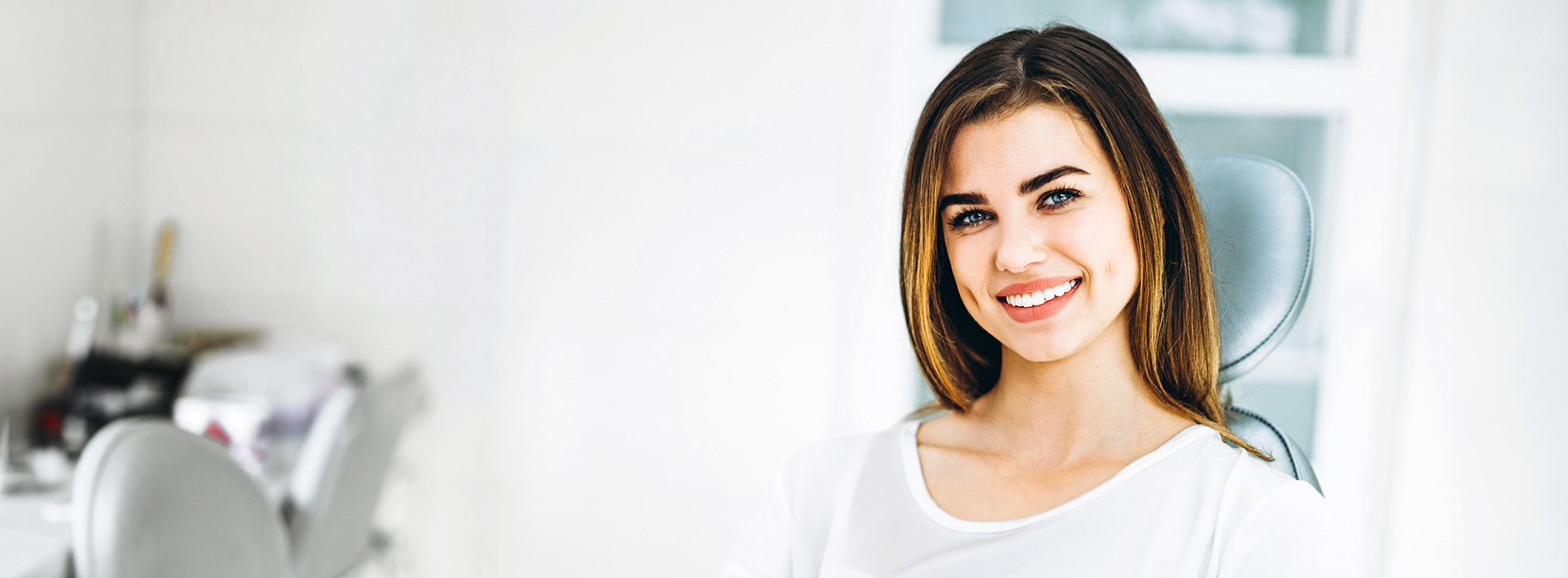 The image shows a woman with short hair, smiling at the camera, wearing a white top and standing in an indoor setting with a minimalist background.