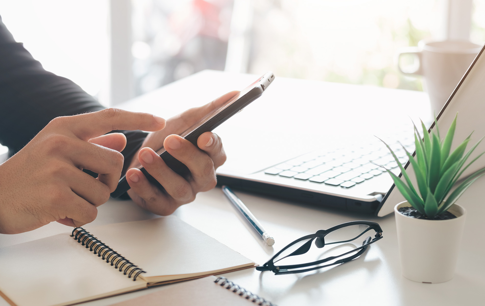 The image shows a person s hands holding a smartphone, with the phone displaying text. The person is seated at a desk with various items, including a laptop, notepad, and pen. In front of the person are a pair of glasses and a small potted plant. The setting appears to be an office environment.