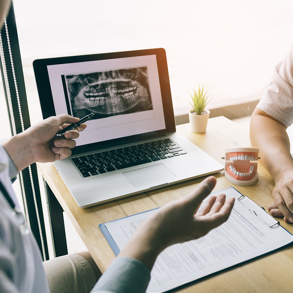 A dental professional is showing a patient an X-ray of their teeth, with both seated at a table in a modern office setting.