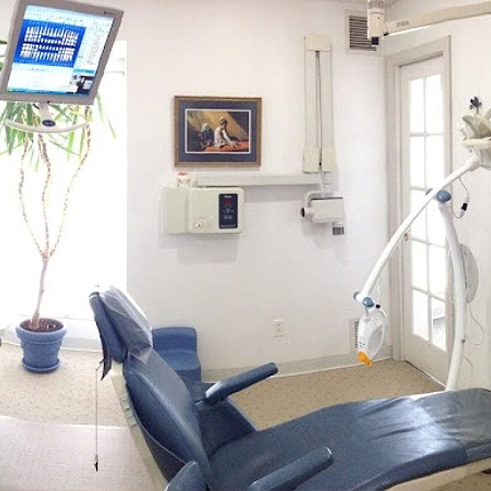 The image shows an interior space, likely a dental or medical office, featuring a dental chair with an adjustable headrest and a computer monitor displaying what appears to be a digital dental X-ray image. In the background, there is a wall-mounted television screen showing a graphical representation of a person s teeth, possibly during a dental procedure or consultation. To the left, a framed picture hangs on the wall, and below it, there is a small table with a book on it. The room has a white color scheme and a window allowing natural light to enter.